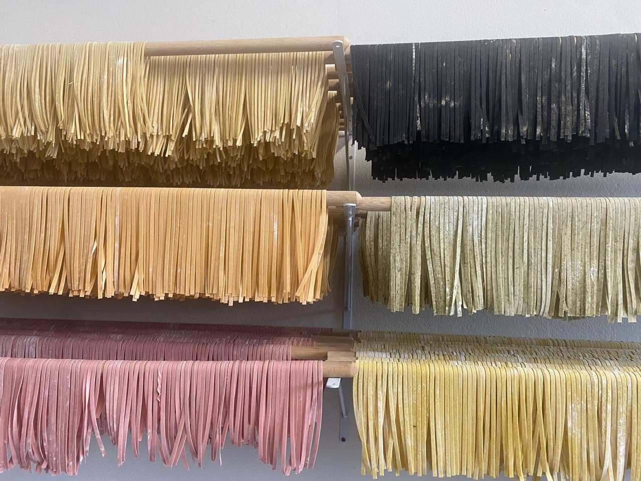 Massas artesanais coloridas secando em varal, do preto ao verde, rosa e amarelo